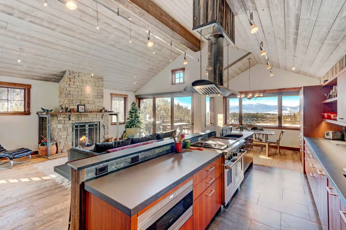 An impressive open living area with a cozy stone fireplace, expansive windows revealing breathtaking mountain views, and an open kitchen layout designed for entertaining and connection. Plush seating creates a comfortable gathering spot for guests.
