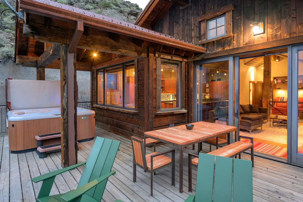 This image features a spacious wooden deck with an inviting hot tub and a dining table set for outdoor meals. The deck overlooks a dramatic rocky landscape, with mountains visible in the distance and a warm, tranquil evening light. The scene reflects both relaxation and the potential for social gatherings, perfectly encapsulating outdoor living in the Rockies.