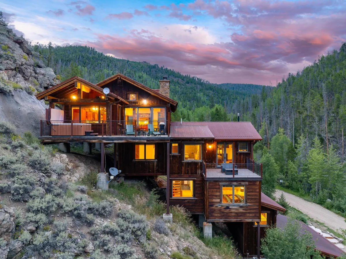 The hero image showcases the chalet's spacious wooden deck overlooking a breathtaking mountain backdrop, with towering pines and a colorful sunset sky. Large windows reflect the tranquil scenery, inviting the outdoors in. The setting evokes warmth and relaxation, hinting at activities like outdoor dining, stargazing, and soaking in the peace of nature.