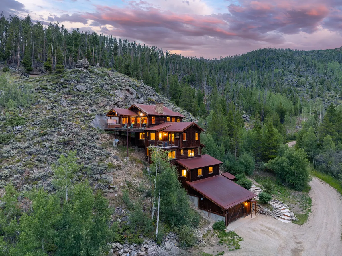 The exterior shot centers on the rustic cabin, set against lush greenery and a stunning mountain backdrop under a glowing sunset sky. The inviting porch and open surroundings highlight the property’s seamless integration into its natural environment, while suggesting direct trail access and space for a variety of outdoor activities.
