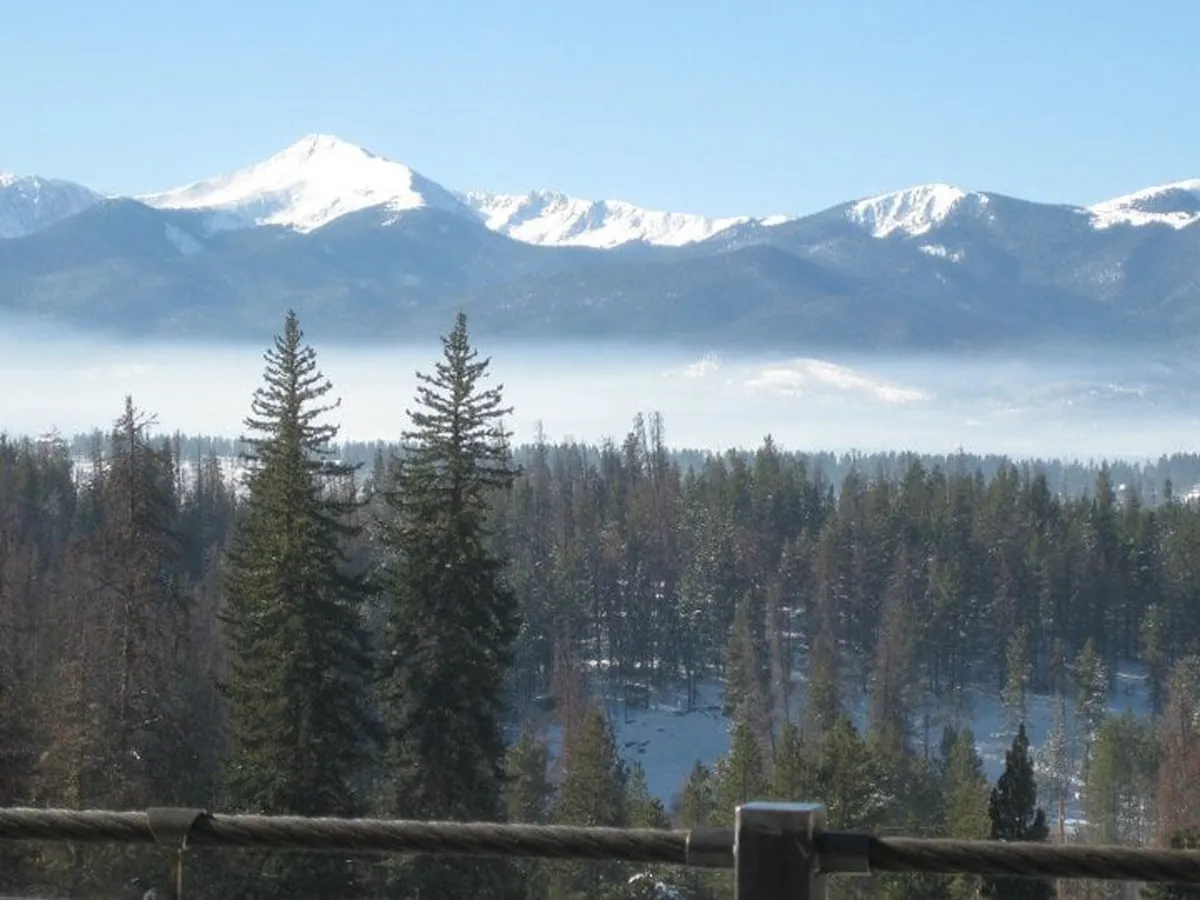 A breathtaking view of snow-capped mountains surrounded by dense evergreen trees, with a misty landscape that creates a sense of tranquility and grandeur. The scene is perfect for outdoor adventurers and those seeking a peaceful retreat in nature.