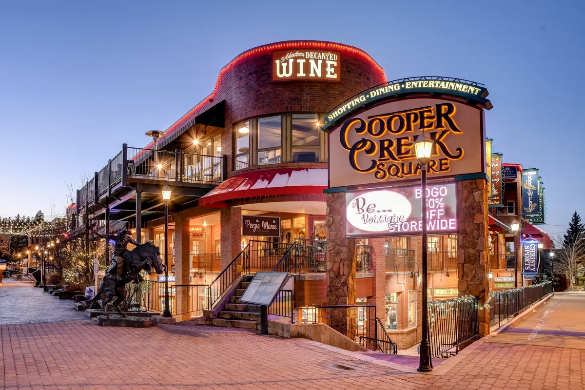 This image shows the vibrant exterior of Cooper Creek Square, featuring the illuminated signage, a wine shop, and surrounding architecture adorned with festive lights. The lively atmosphere is evident with hints of shopping, dining, and entertainment activities taking place.