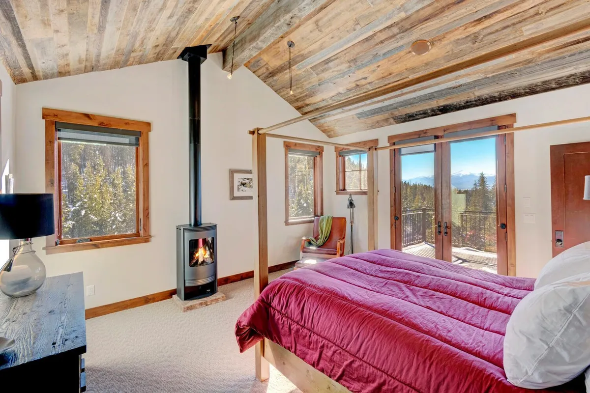 A tranquil bedroom featuring a plush four-poster bed, a cozy wood stove, and large windows framing mountain views. The room exudes warmth and comfort, ideal for restful nights or quiet mornings with nature as your backdrop.