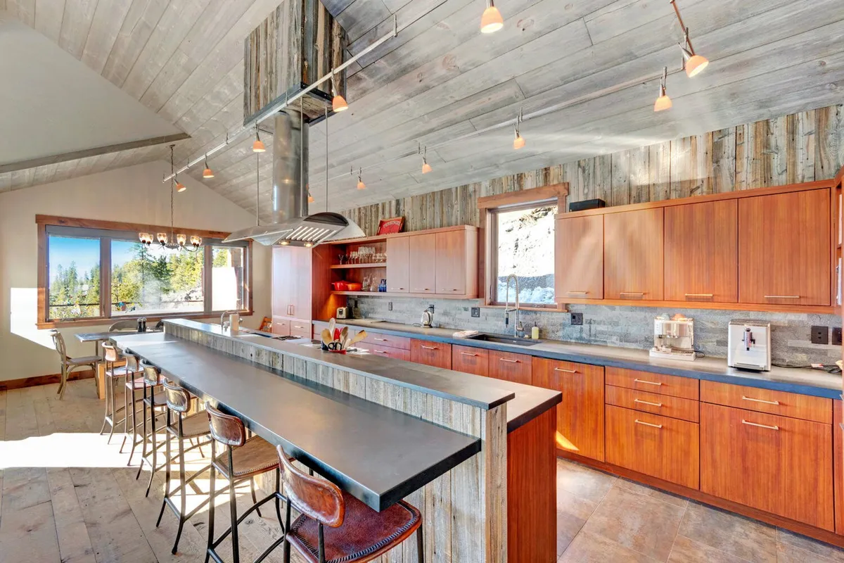 This image features a long kitchen island, wooden cabinets, and large windows that flood the space with light. The layout encourages mingling while preparing meals and highlights the kitchen as a central hub for gathering and entertaining.