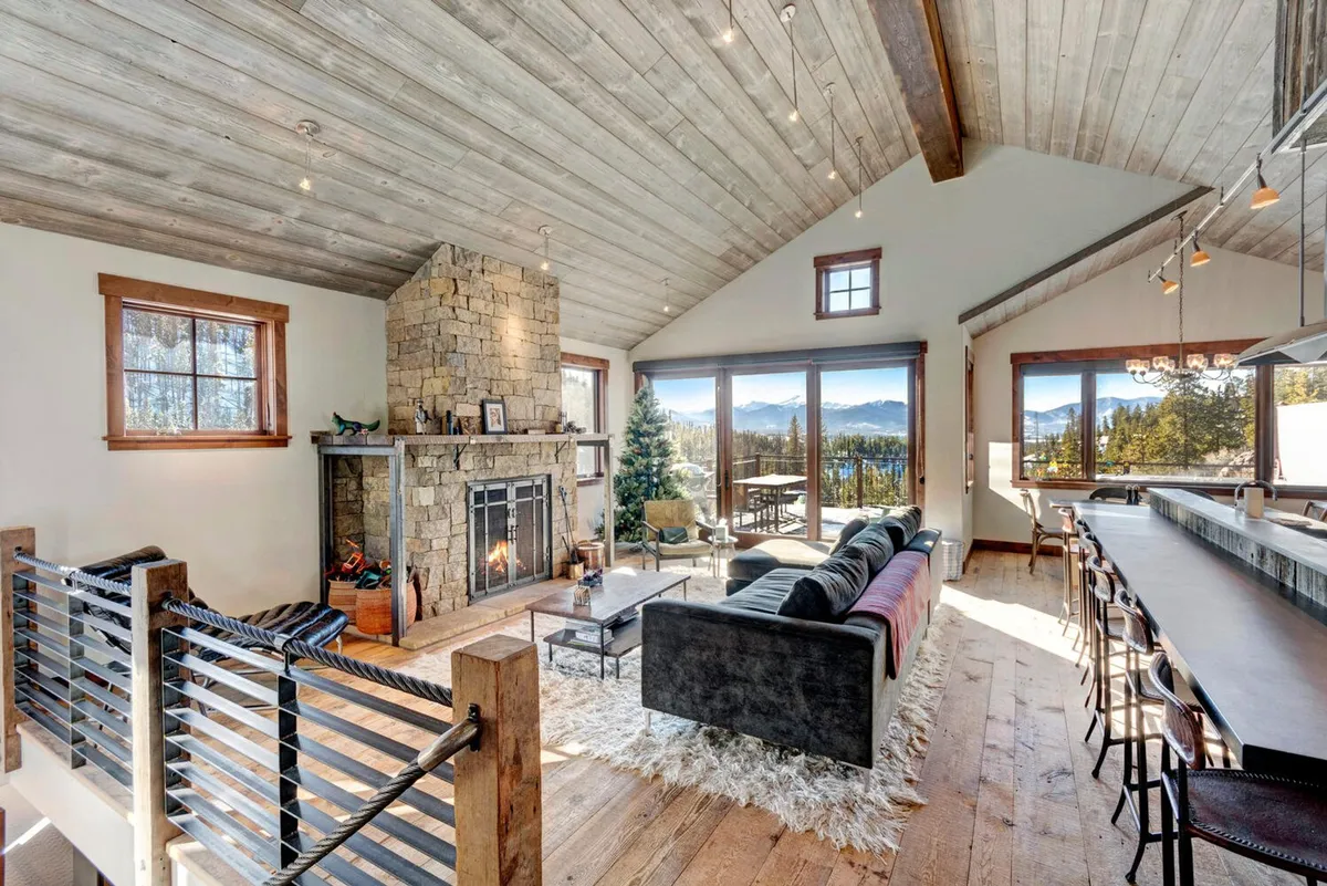 A warm, inviting great room featuring a dramatic stone fireplace as the focal point, with a comfortable sofa arranged to soak in the panoramic mountain views through expansive floor-to-ceiling windows. The open layout seamlessly connects to a modern kitchen island, creating a spacious setting for gatherings, relaxation, and entertainment.