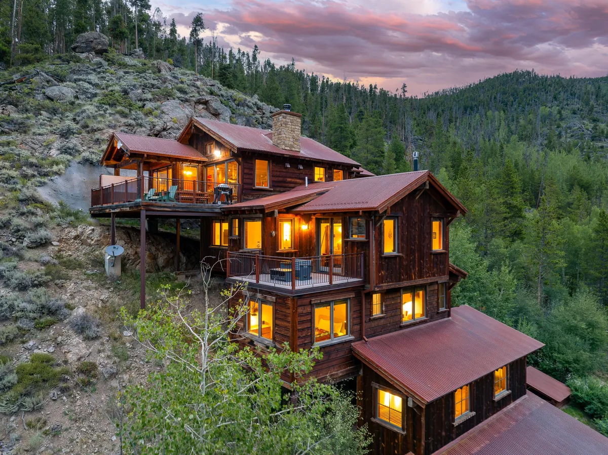 The rustic wooden exterior of the chalet is illuminated by glowing windows, set amid a peaceful natural landscape. The deck offers a prominent gathering space for families to watch sunsets, stargaze, or recount the day's adventures. The image conveys warmth and tranquility, inviting guests to imagine evenings spent together after exploring the outdoors.