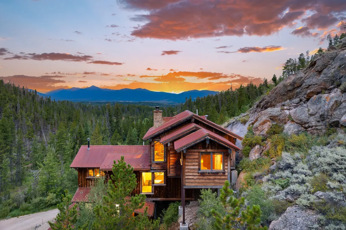 This exterior image highlights the Cliffside Luxury Chalet set against a dramatic mountain range beneath a colorful sunset sky. The house appears nestled in nature, with the surrounding landscape suggesting direct access to hiking trails and the bordering National Forest. The warm glow from within the home hints at comfort and family togetherness after a day of outdoor adventure.